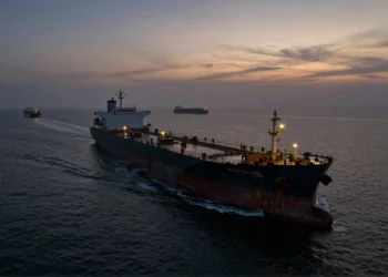 Superpetrolero oxidado y anónimo navega solo al atardecer por un estrecho marítimo concurrido, sin bandera visible