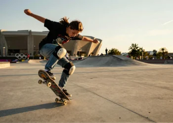 Julia Benedetti ejecutando un kickflip en una plaza de concreto urbano con el estadio olímpico desenfocado al fondo