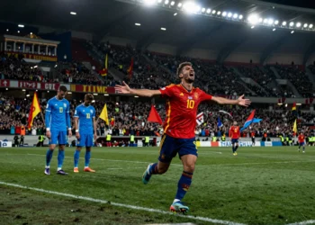 Futbolista español celebra con los brazos abiertos ante dos defensas uruguayos cabizbajo en un estadio mundialista nocturno