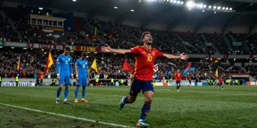 Futbolista español celebra con los brazos abiertos ante dos defensas uruguayos cabizbajo en un estadio mundialista nocturno