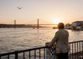 Hombre contemplando el estuario del río Tajo desde el paseo marítimo de Lisboa al atardecer, con el puente al fondo