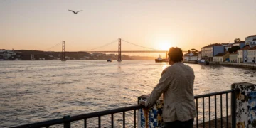 Hombre contemplando el estuario del río Tajo desde el paseo marítimo de Lisboa al atardecer, con el puente al fondo
