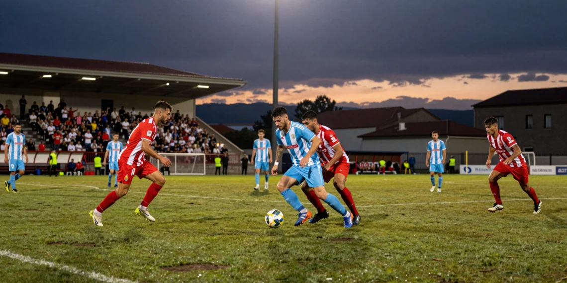 Duelo de fútbol en A Lomba bajo los focos nocturnos entre jugadores con camisetas a rayas rojas y blancas y azul celeste