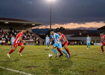 Duelo de fútbol en A Lomba bajo los focos nocturnos entre jugadores con camisetas a rayas rojas y blancas y azul celeste