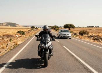 Motociclista con casco homologado en carretera rural española de dos carriles, con un coche adelante bajo el sol mediterráneo