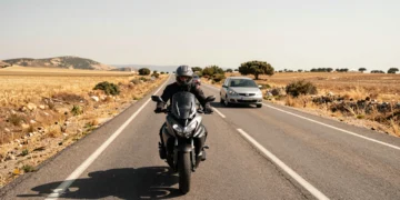Motociclista con casco homologado en carretera rural española de dos carriles, con un coche adelante bajo el sol mediterráneo