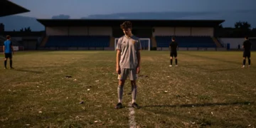 Futbolista joven solo en campo vacío al atardecer, con tres figuras distantes representando clubes en negociación