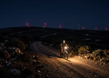 Ciclista solitario iluminando una pista de tierra nocturna en Terres de l'Ebre con luces de molinos al fondo