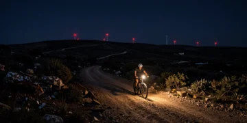 Ciclista solitario iluminando una pista de tierra nocturna en Terres de l'Ebre con luces de molinos al fondo