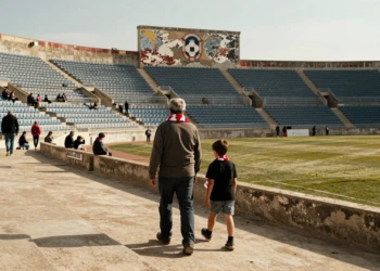 Aficionado mayor y niño con bufandas rojiblancas caminan hacia un estadio semivacío bajo la luz del sol en Murcia