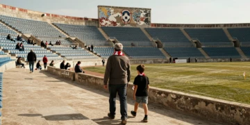 Aficionado mayor y niño con bufandas rojiblancas caminan hacia un estadio semivacío bajo la luz del sol en Murcia