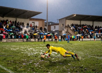 Portero en equipación amarilla se lanza a detener un balón mojado bajo los focos de un estadio municipal catalán al atardecer