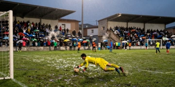 Portero en equipación amarilla se lanza a detener un balón mojado bajo los focos de un estadio municipal catalán al atardecer