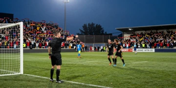 Árbitro señalando penalti en estadio de fútbol de Primera Federación española al atardecer con aficionados en las gradas