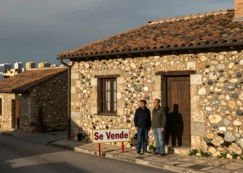 Pareja inspecciona vivienda tradicional en venta en pueblo de Teruel, contraste con skyline urbano de Madrid al fondo